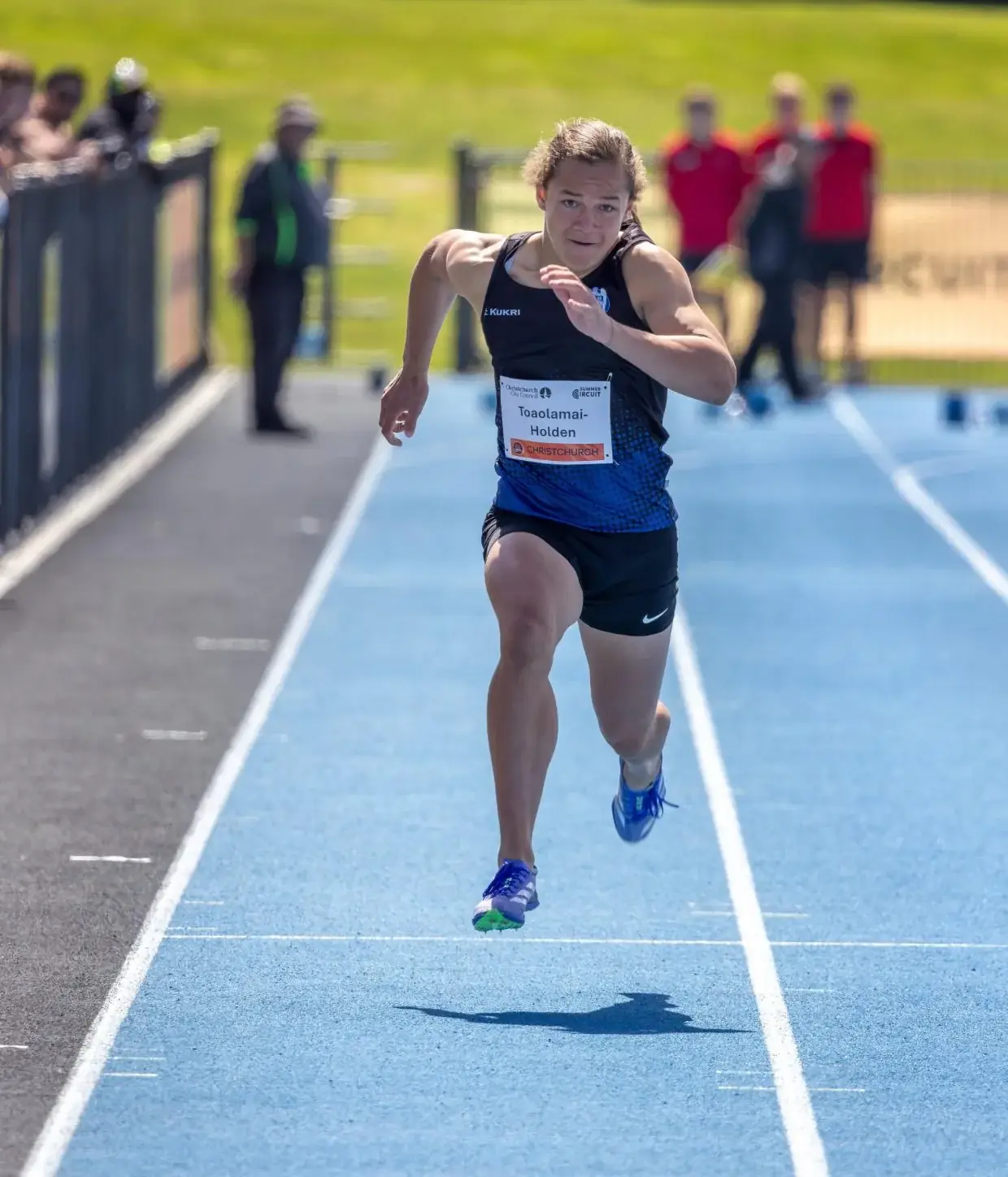Vernie, junior sprinter, running on a blue track in black and blue racing kit with an intense focused expression